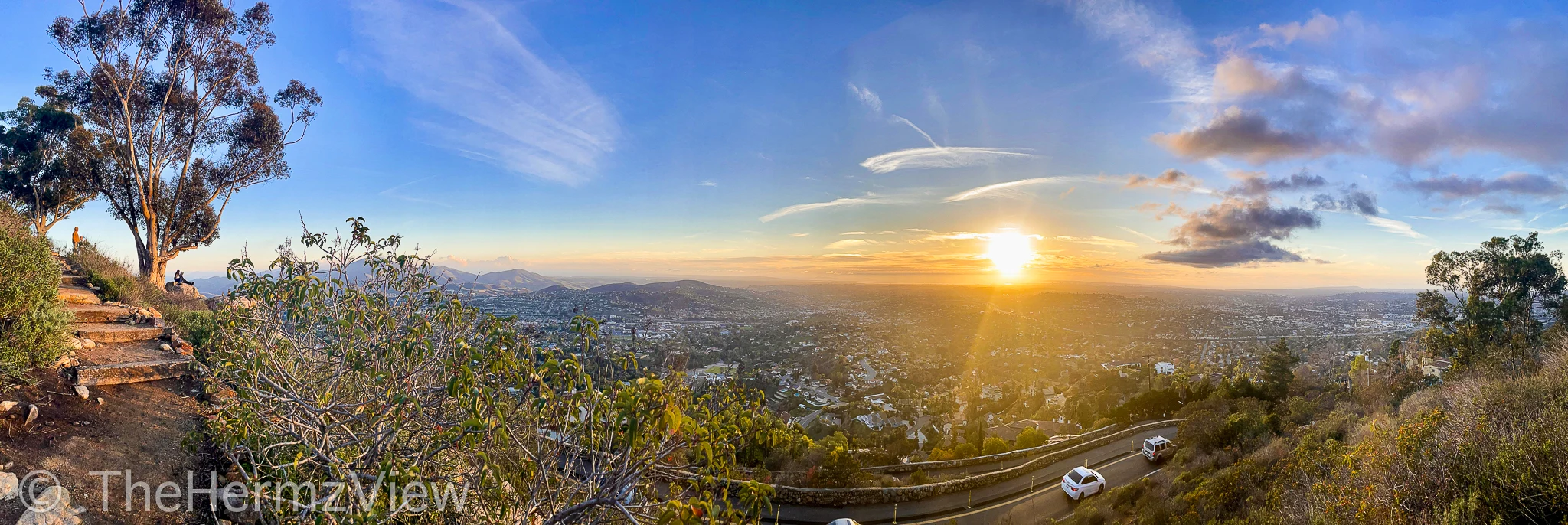 Panoramic sunset view looking down from a hillside path onto a vast valley with a winding road below and bright sun low on the horizon.