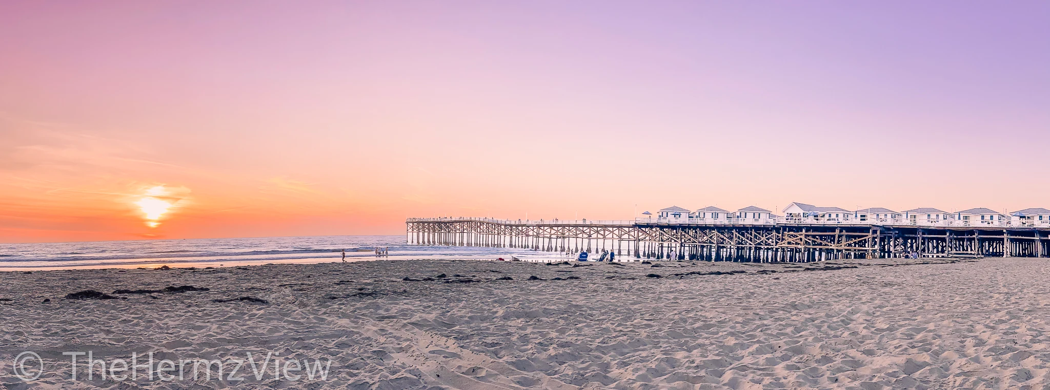 Wide panoramic view of a sandy beach and wooden pier extending into the sea under a sunset sky gradient of orange and soft purple.