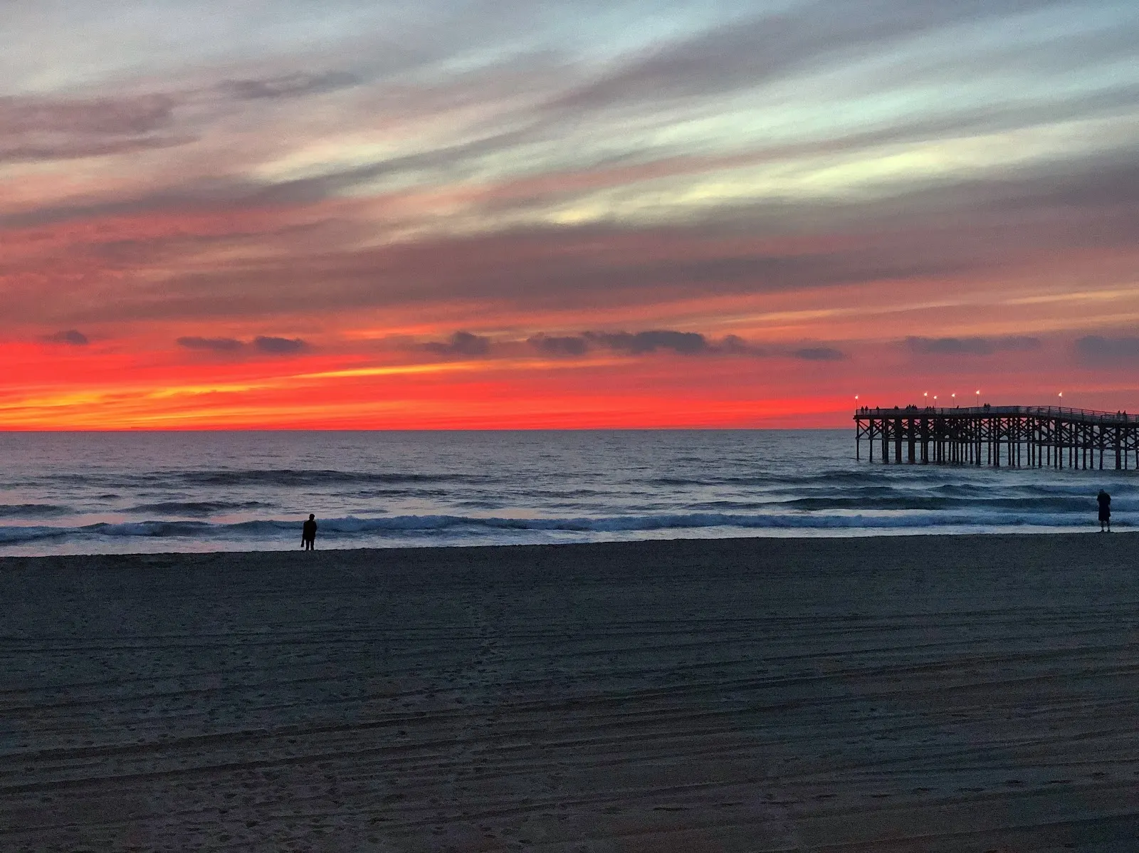 Scarlet sunset over the Pacific Ocean with a pier and two people silhouetted along the beach.