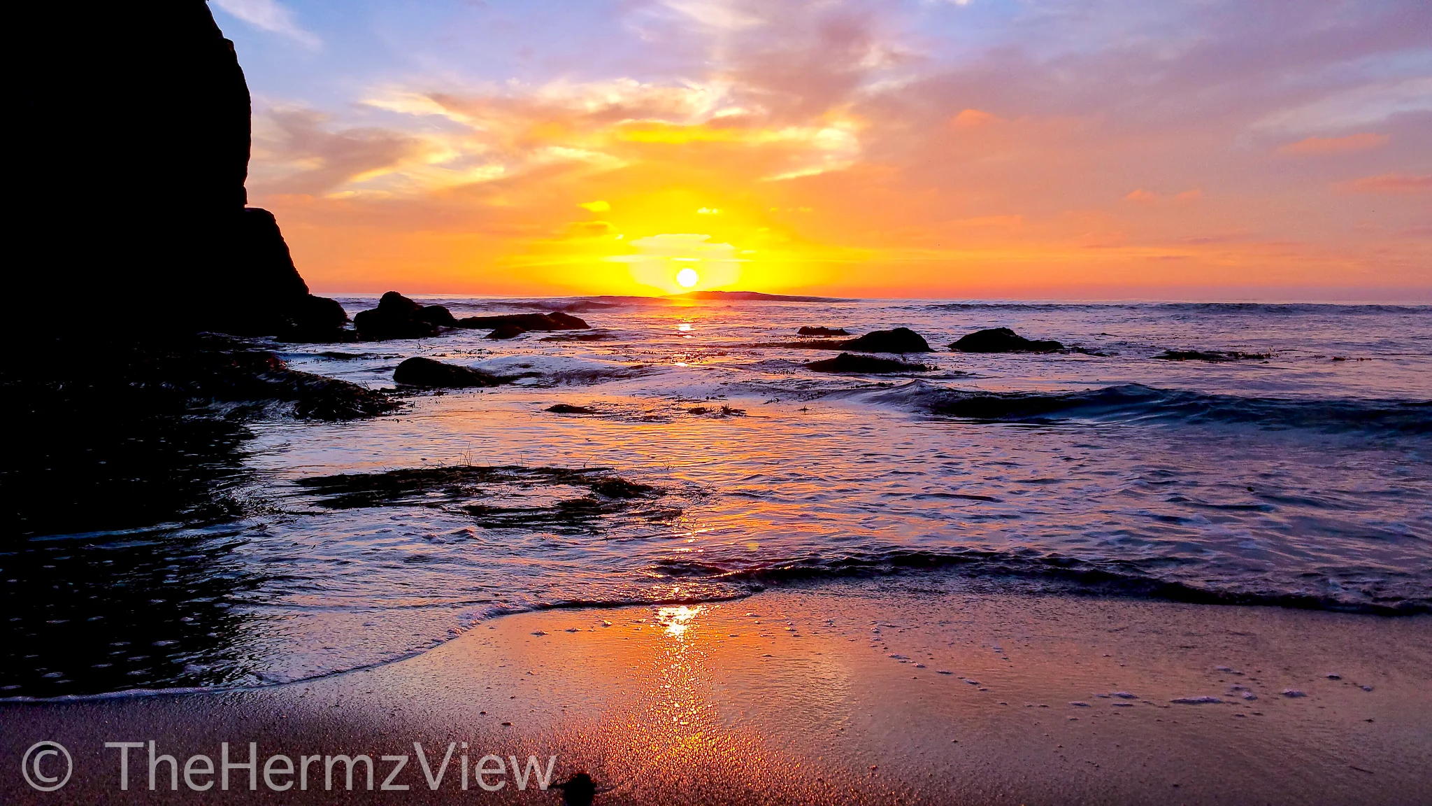 A sunset over the ocean with golden sun rays reflecting on wet sand and waves framed by dark coastal rocks.