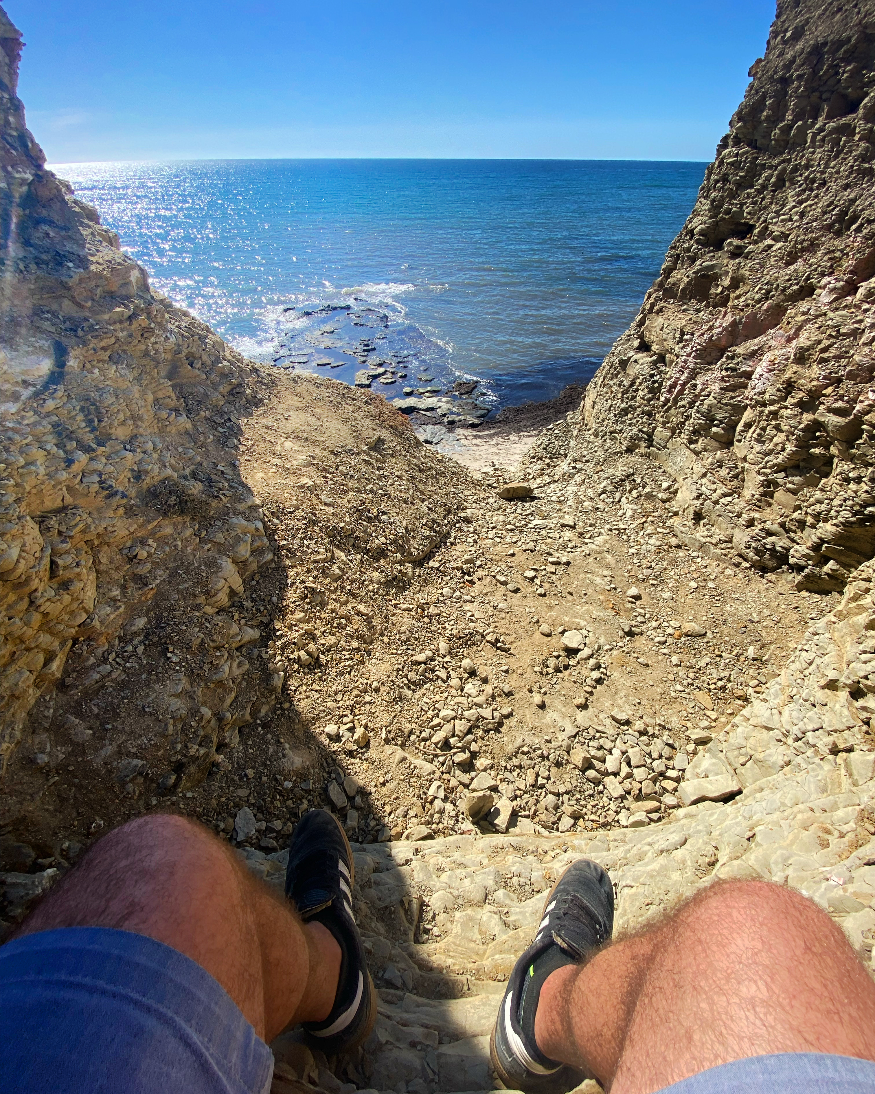 First-person view looking down a steep rocky cliff path framing a bright blue ocean with sparkling sunlight and tide pools.