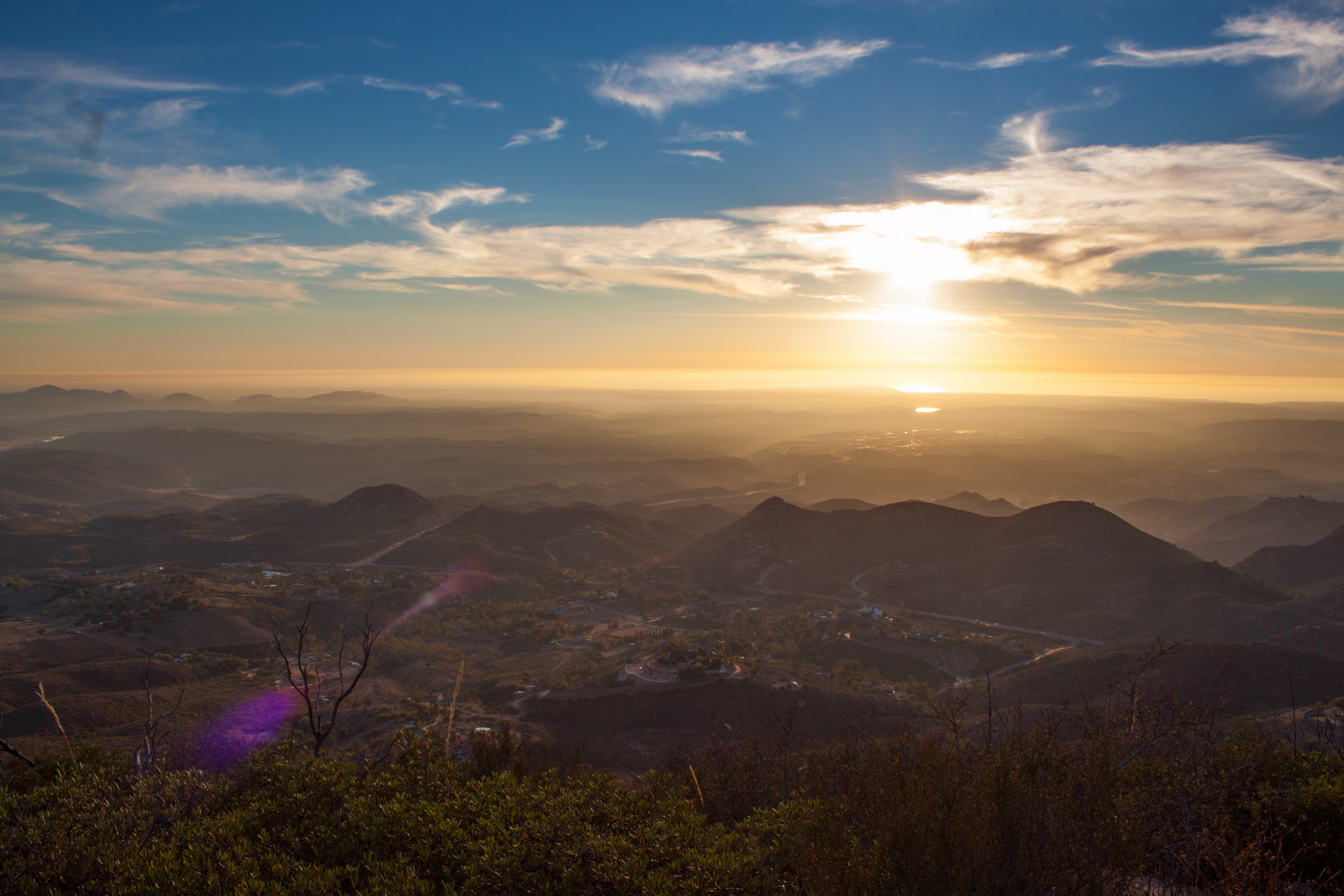 Wide view from a high ridge looking over layered mountain slopes and hazy valleys toward a bright sun low on the horizon.