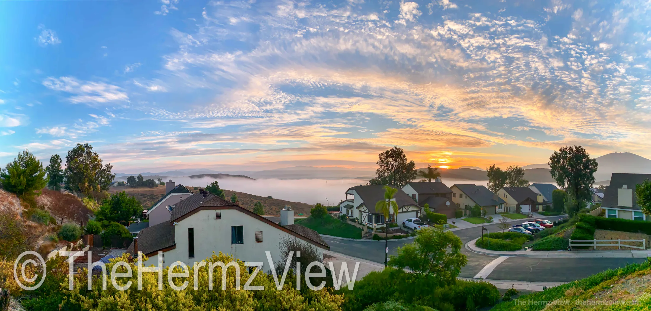 Panoramic view of a suburban neighborhood overlooking a fog-filled valley during sunrise under wispy clouds.