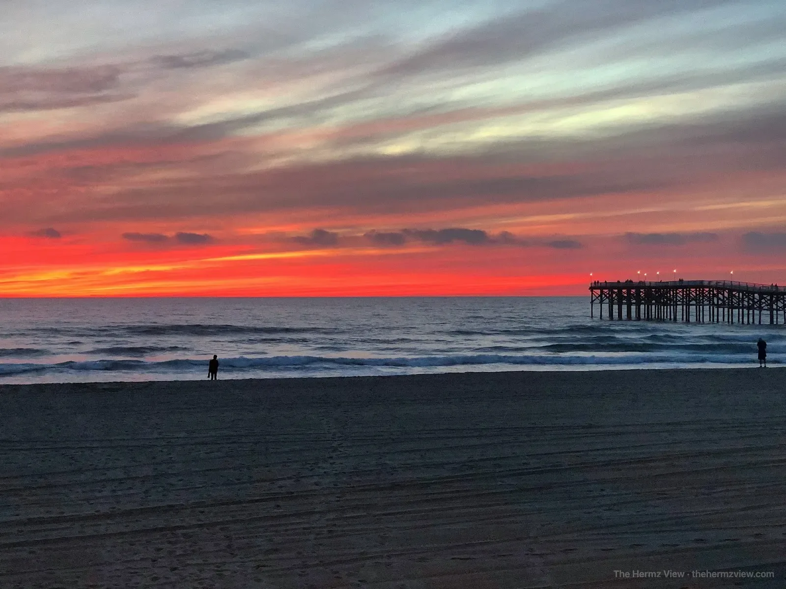 Scarlet sunset over the Pacific Ocean with a pier and two people silhouetted along the beach.