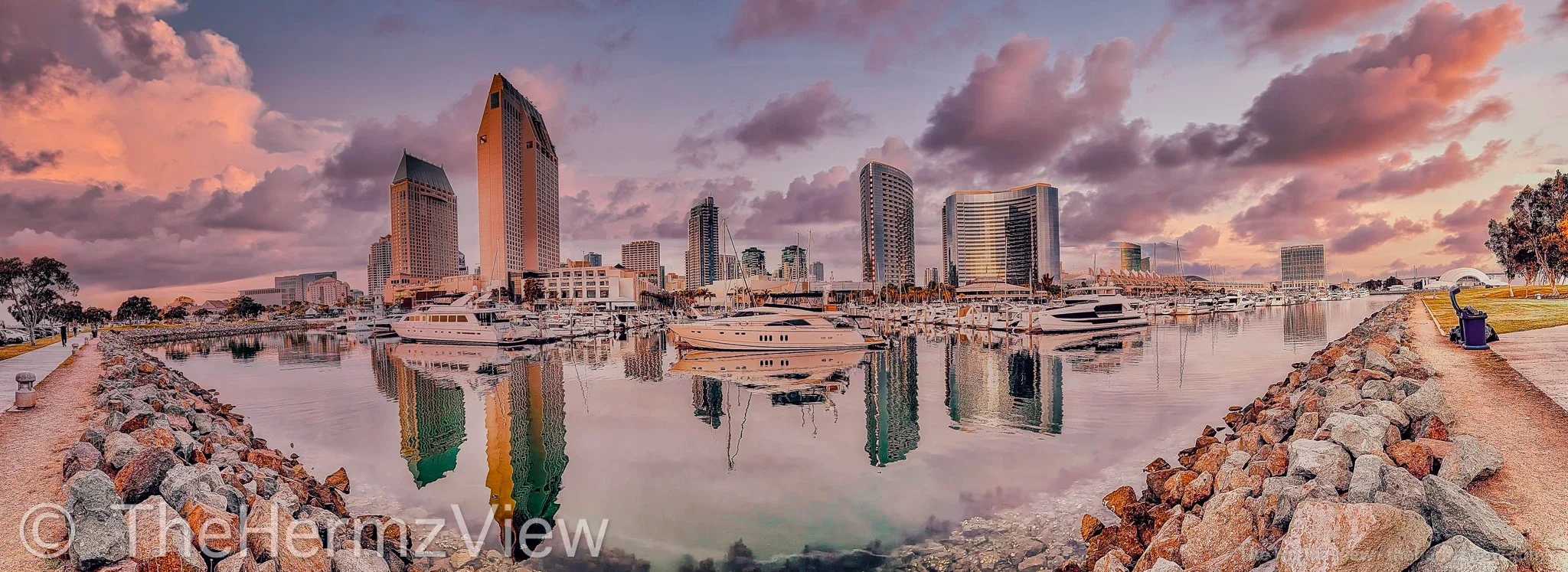 Panoramic wide-angle view of a city skyline across a marina with calm water reflections, rocky shorelines, and pink twilight clouds.