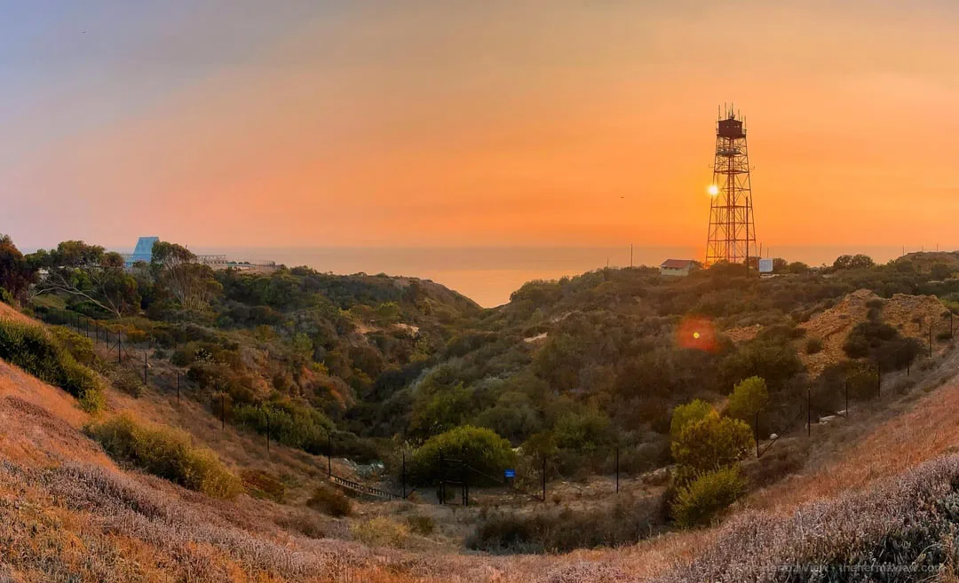 A tall steel lattice tower stands on a coastal ridge backlit by a hazy orange sunset over a brush-covered canyon leading to the ocean horizon.