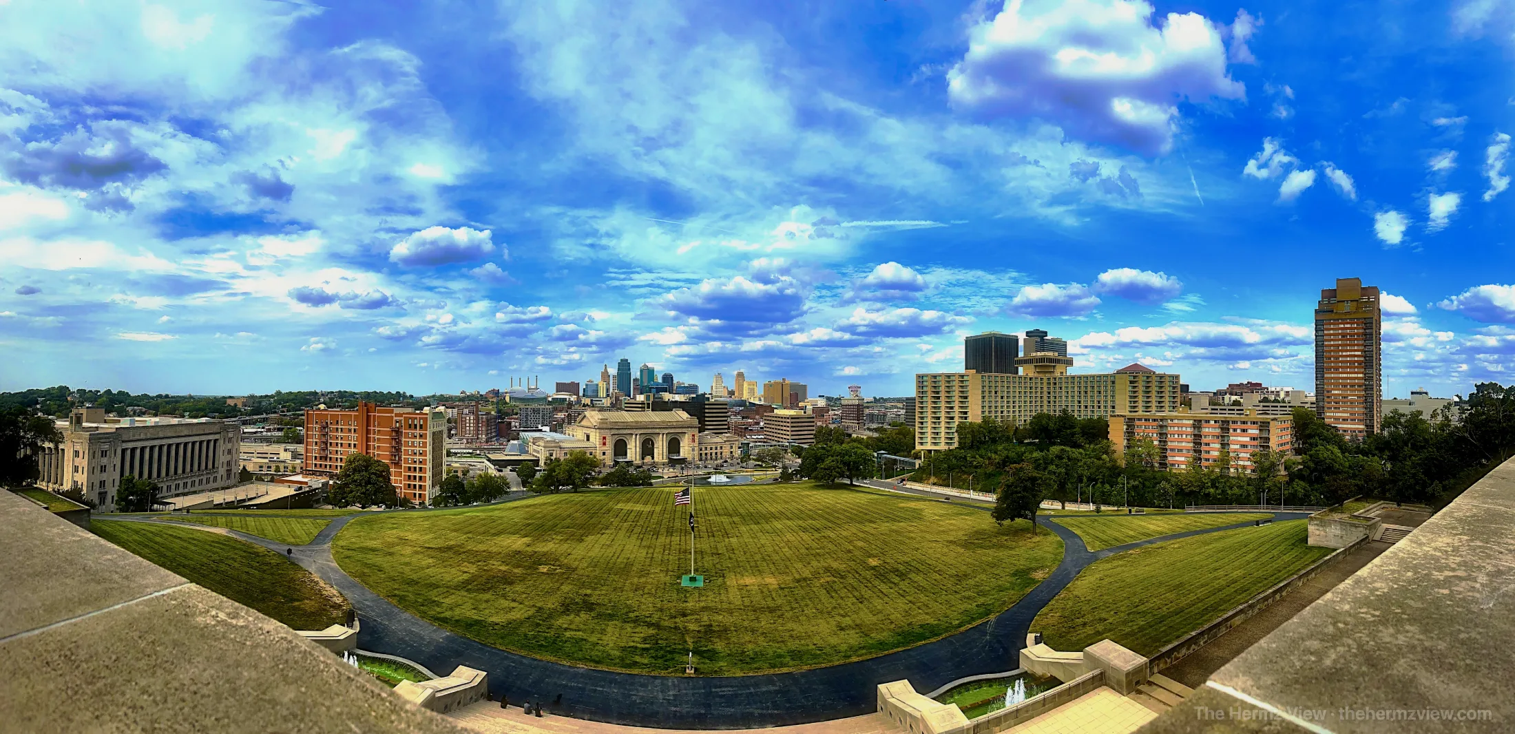 Panoramic wide view of a city skyline framing an expansive green lawn with stone steps in the foreground under a vibrant blue sky filled with white clouds.