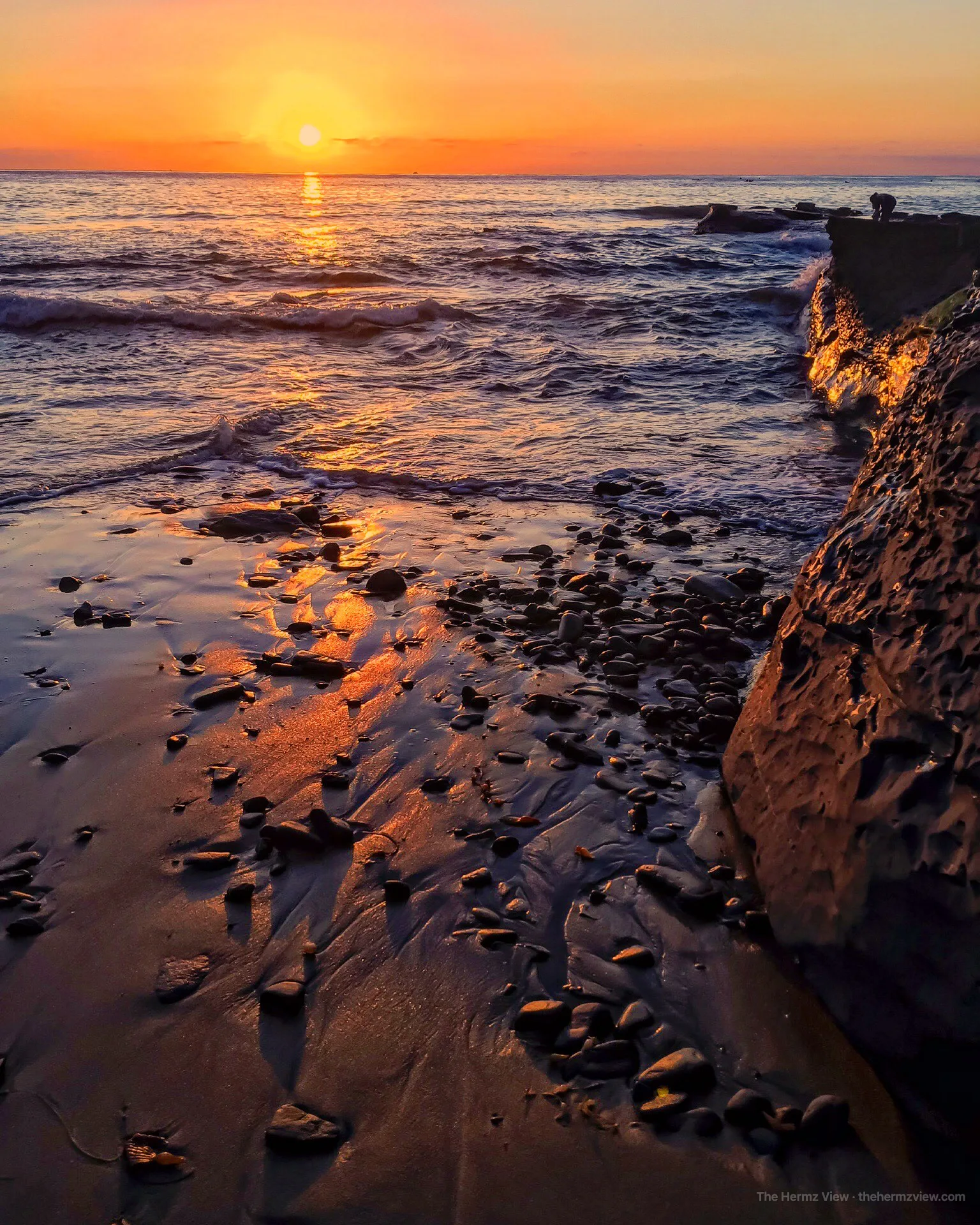 Bright orange sunset over the ocean reflecting on wet sand and rocks along a coastal shore.
