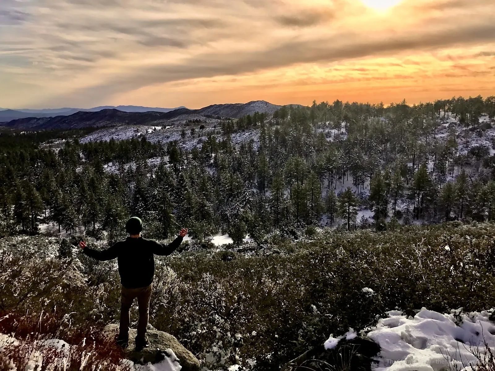 Person overlooking a snow-covered mountain forest beneath a warm winter sunset.
