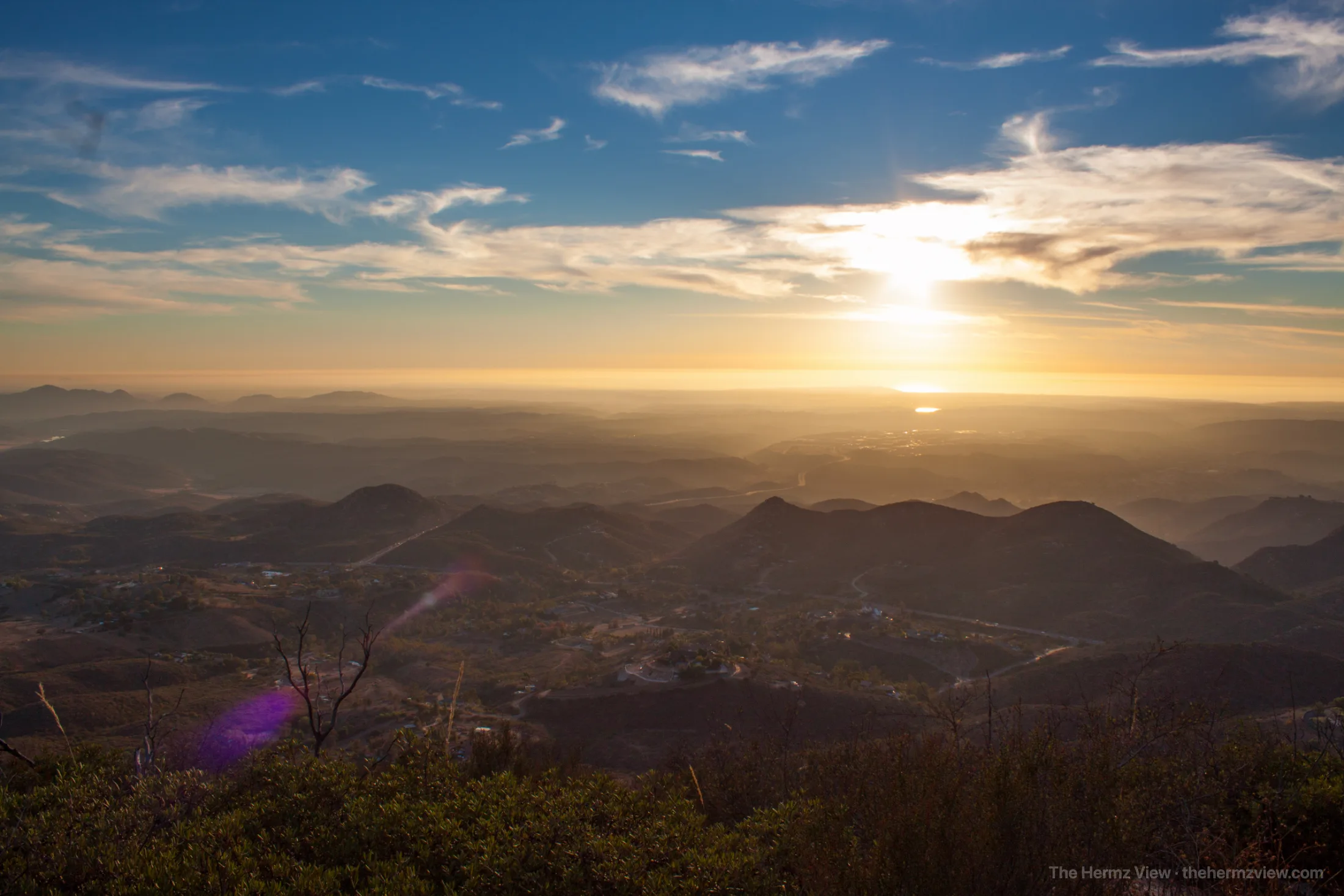 Wide view from a high ridge looking over layered mountain slopes and hazy valleys toward a bright sun low on the horizon.