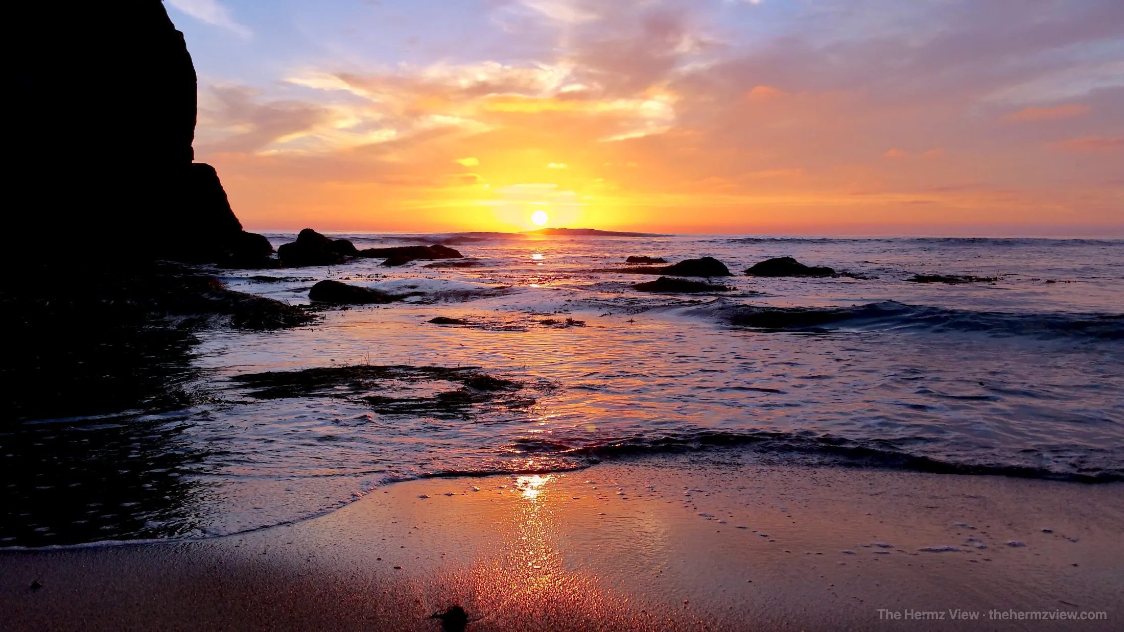 Sunset over a calm ocean beach with dark silhouetted sea cliffs on the left, scattered rocks in shallow water, and golden light reflected on wet sand.