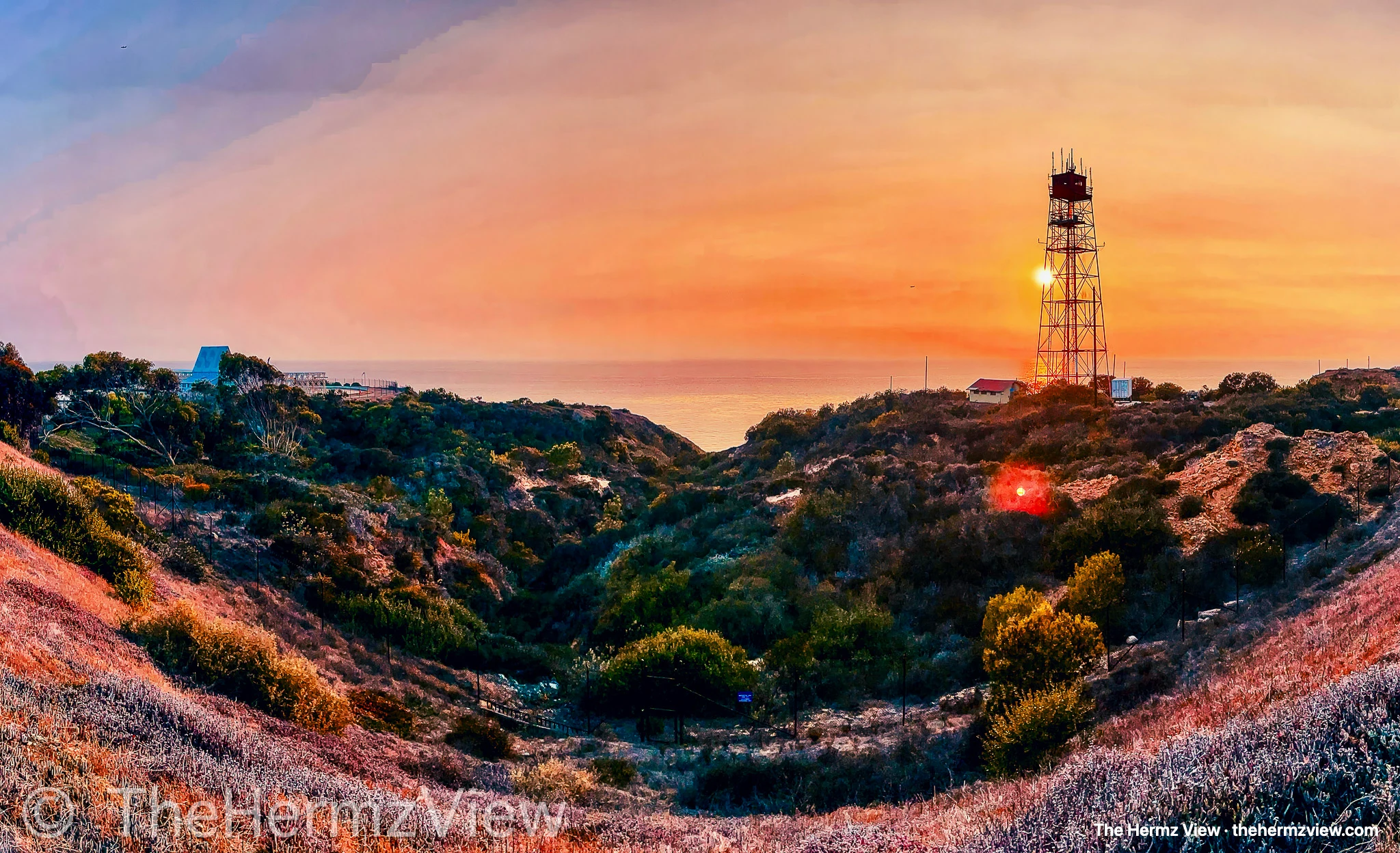 A wide coastal canyon filled with green shrubbery leading to the ocean under a warm orange sunset sky, with a tall metallic lattice tower standing on the right ridge.