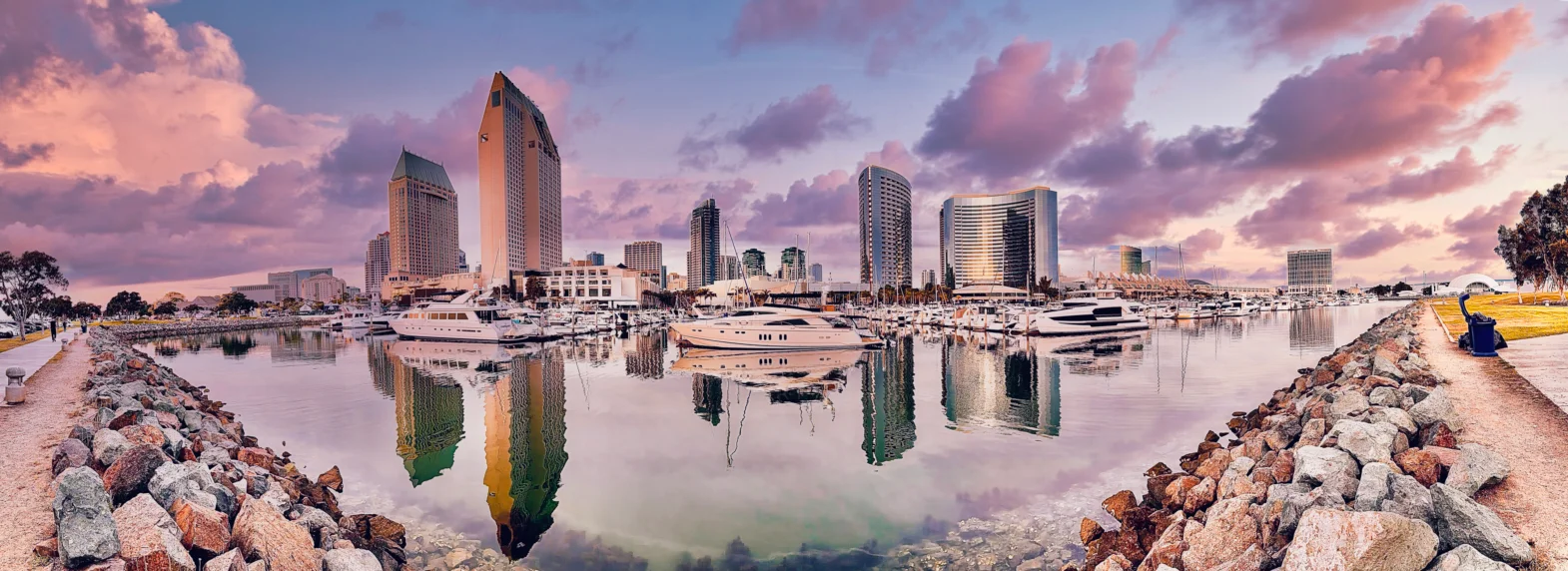 Panoramic view of a city harbor lined with high-rise buildings and docked yachts, framed by rocky shorelines with soft pink and purple clouds reflecting in calm water.
