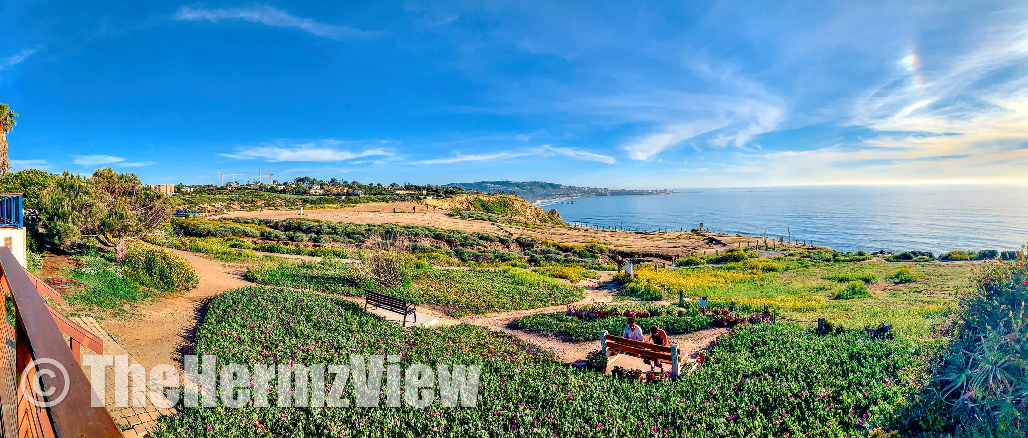 Panoramic vista of green coastal bluffs, dirt pathways with wooden benches, and a wide blue ocean under a bright sky.