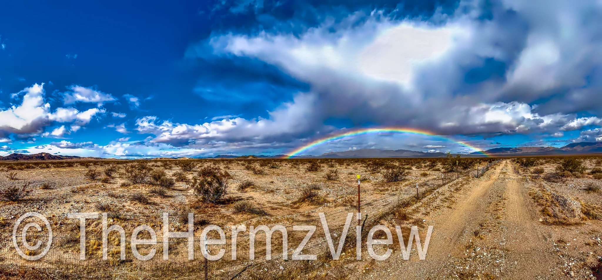 A full rainbow spans the horizon over an open desert plain with a dirt track and wire fence on the right side beneath storm clouds.