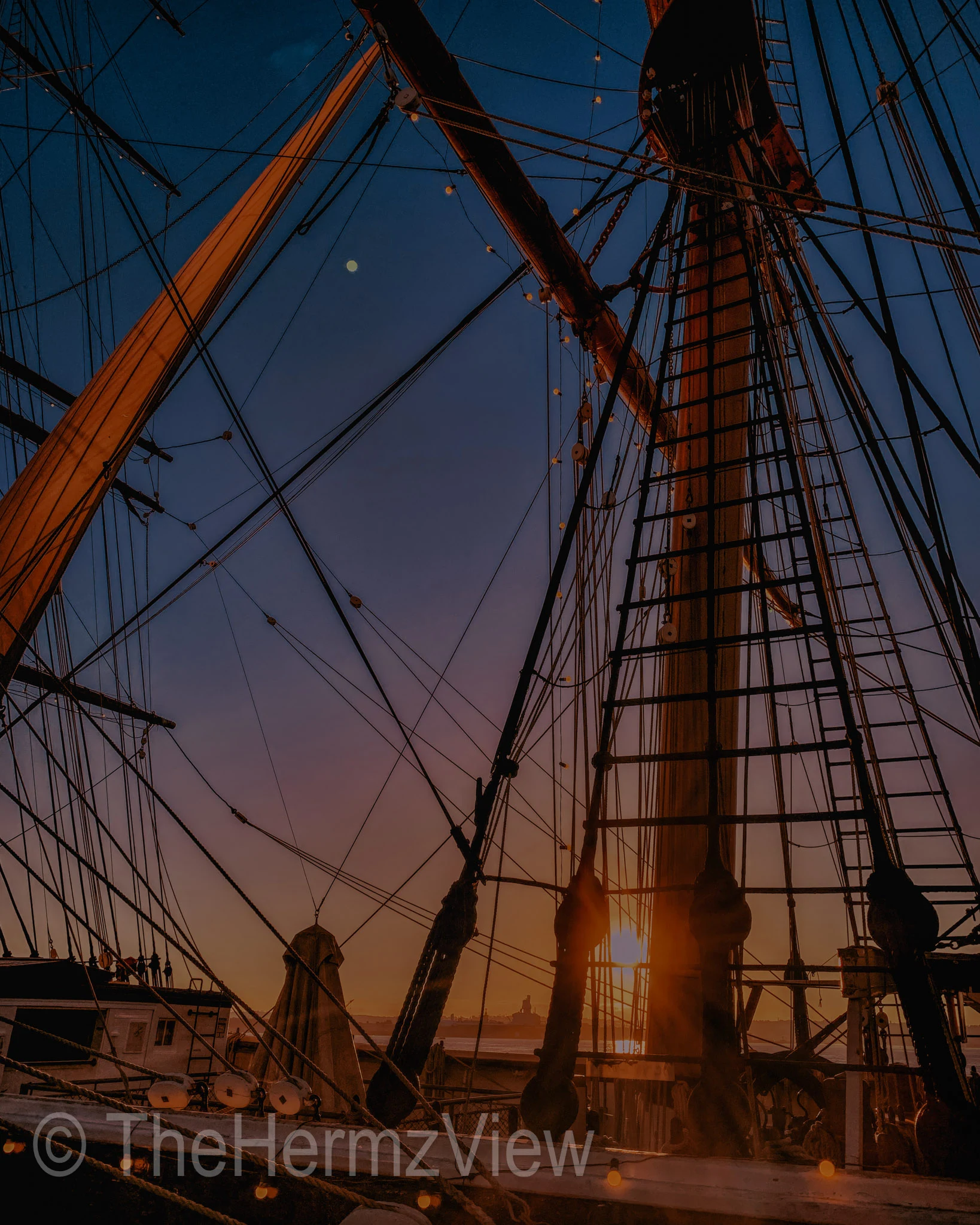Intricate wooden ship masts, ropes, and ratlines silhouetted against a gradient sunset sky fading from dark blue to warm orange.