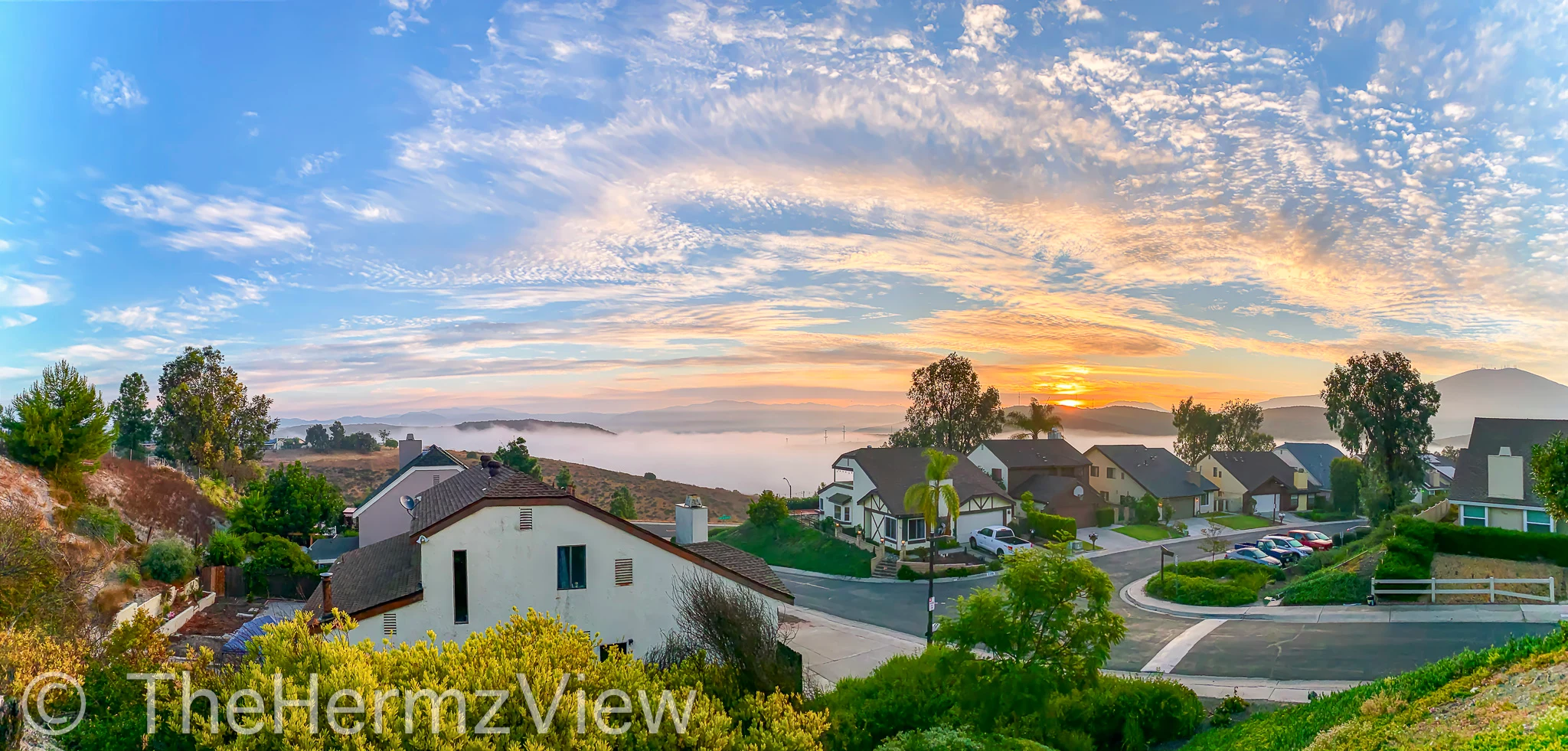 Panoramic view of a hillside suburban neighborhood overlooking a valley covered in low fog under a cloud-filled sky.