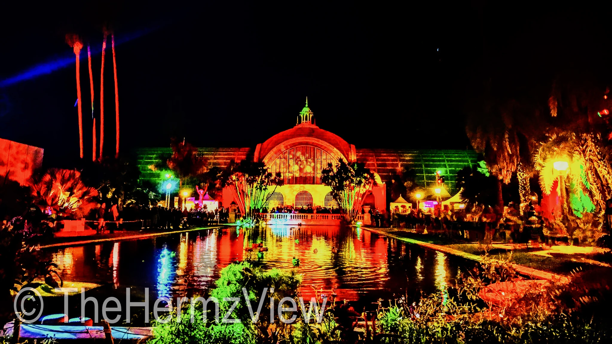 A large domed structure and surrounding palm trees bathed in vivid red and green lights, reflecting across a wide rectangular pond at night.