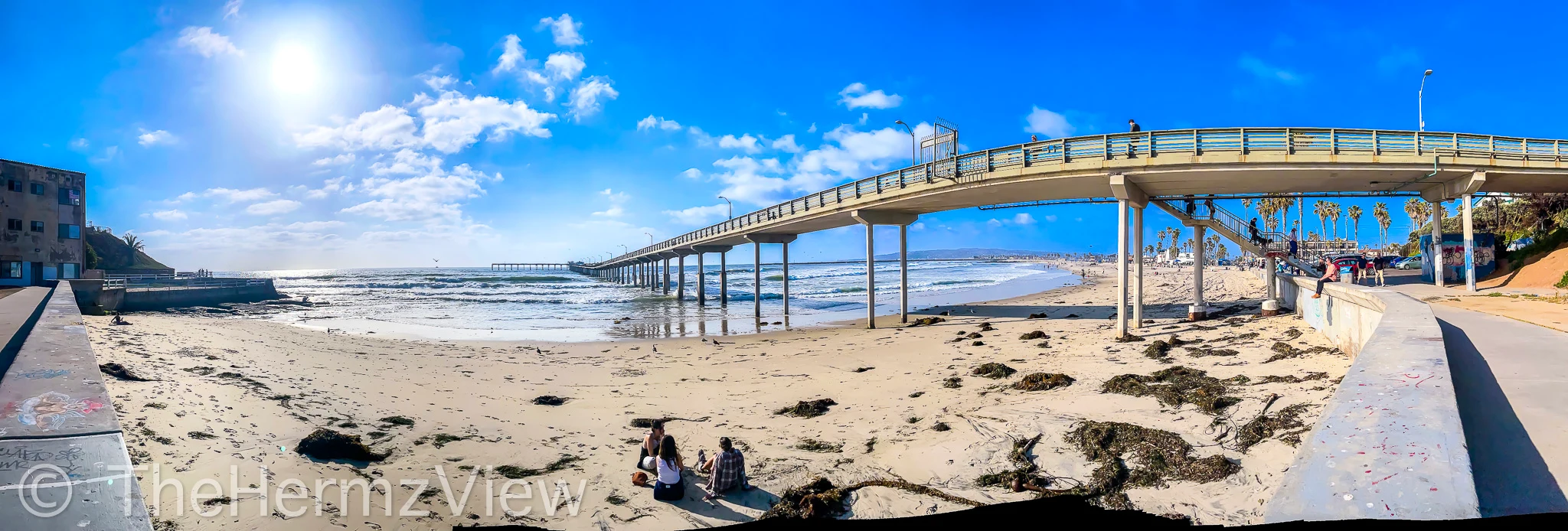 Wide panoramic view of a sandy beach strewn with kelp, framed by a concrete pier stretching out into ocean waves under a bright, clear sun.