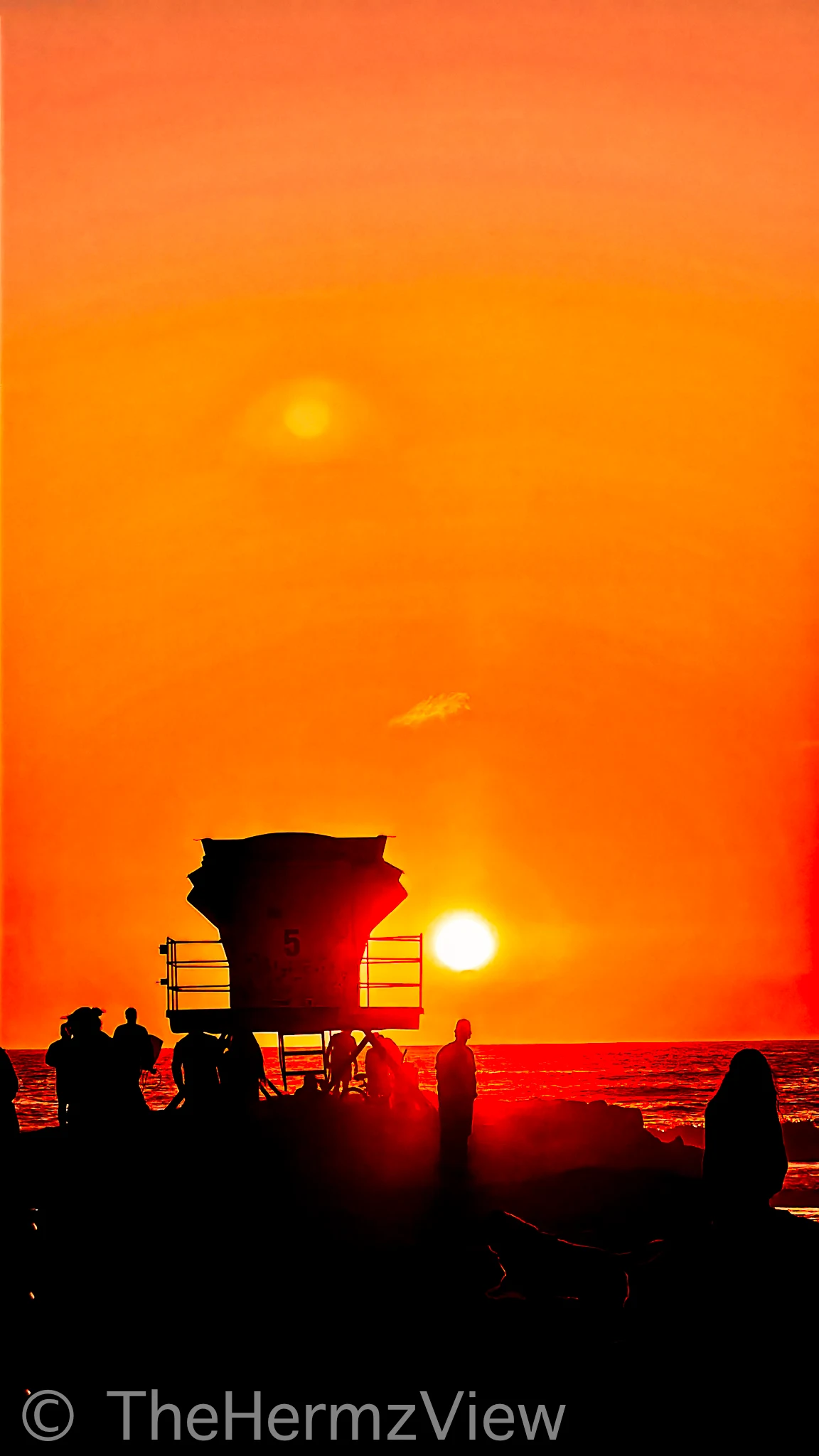 Silhouetted lifeguard tower number 5 and figures along a beach beneath a glowing orange sunset sky.