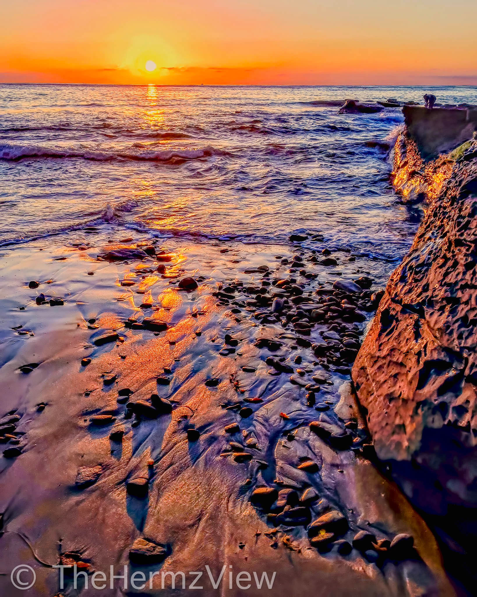 Bright orange sunset over ocean waves reflecting onto wet sandy shoreline with scattered pebbles and rugged cliffs on the right.