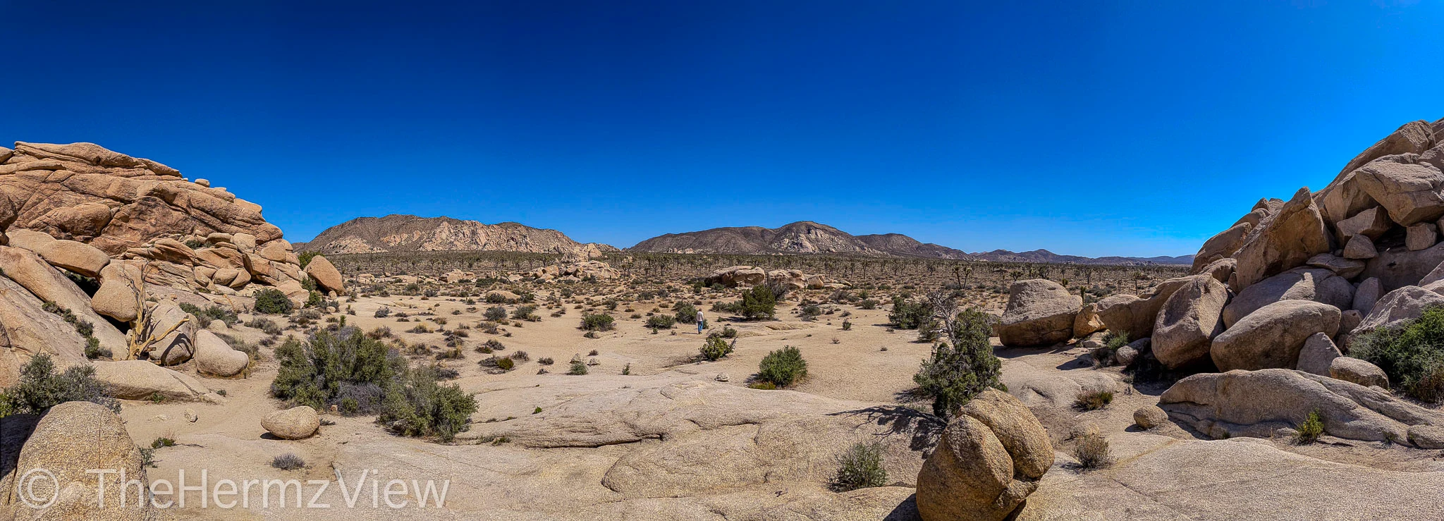 A wide panoramic view of a desert landscape surrounded by large granite boulder formations under a clear blue sky.