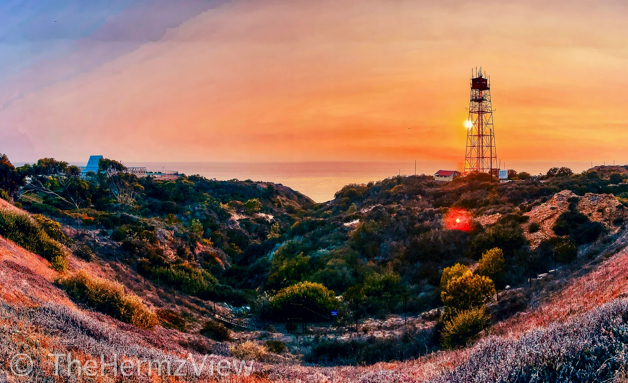 A wide coastal canyon filled with green shrubbery leading to the ocean under a warm orange sunset sky, with a tall metallic lattice tower standing on the right ridge.