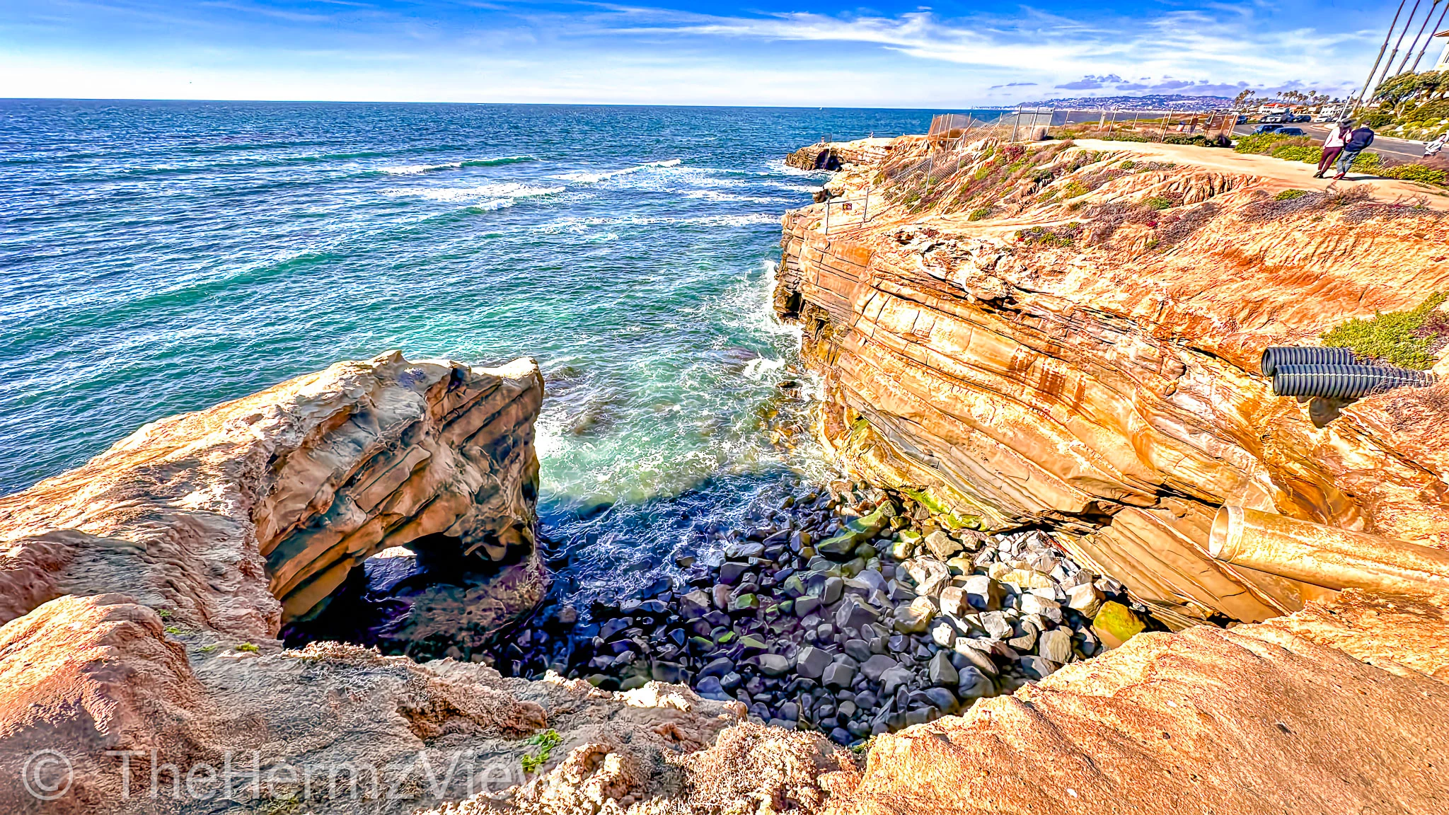 High-angle view of layered coastal sandstone cliffs meeting turquoise ocean water near a natural rock arch and rocky cove.