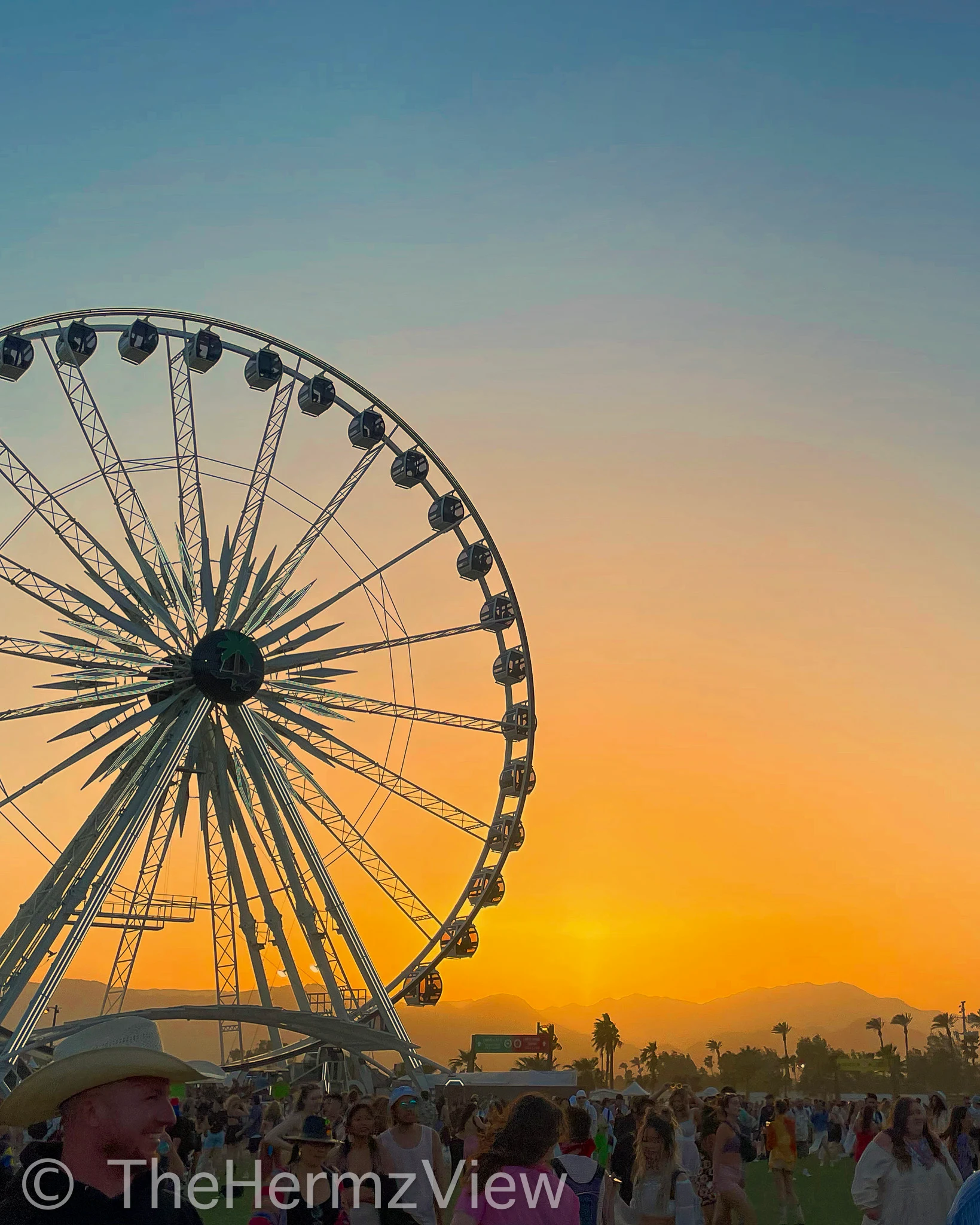 A large Ferris wheel silhouetted against a vibrant orange and blue sunset sky, with distant mountains and a crowd of people below.