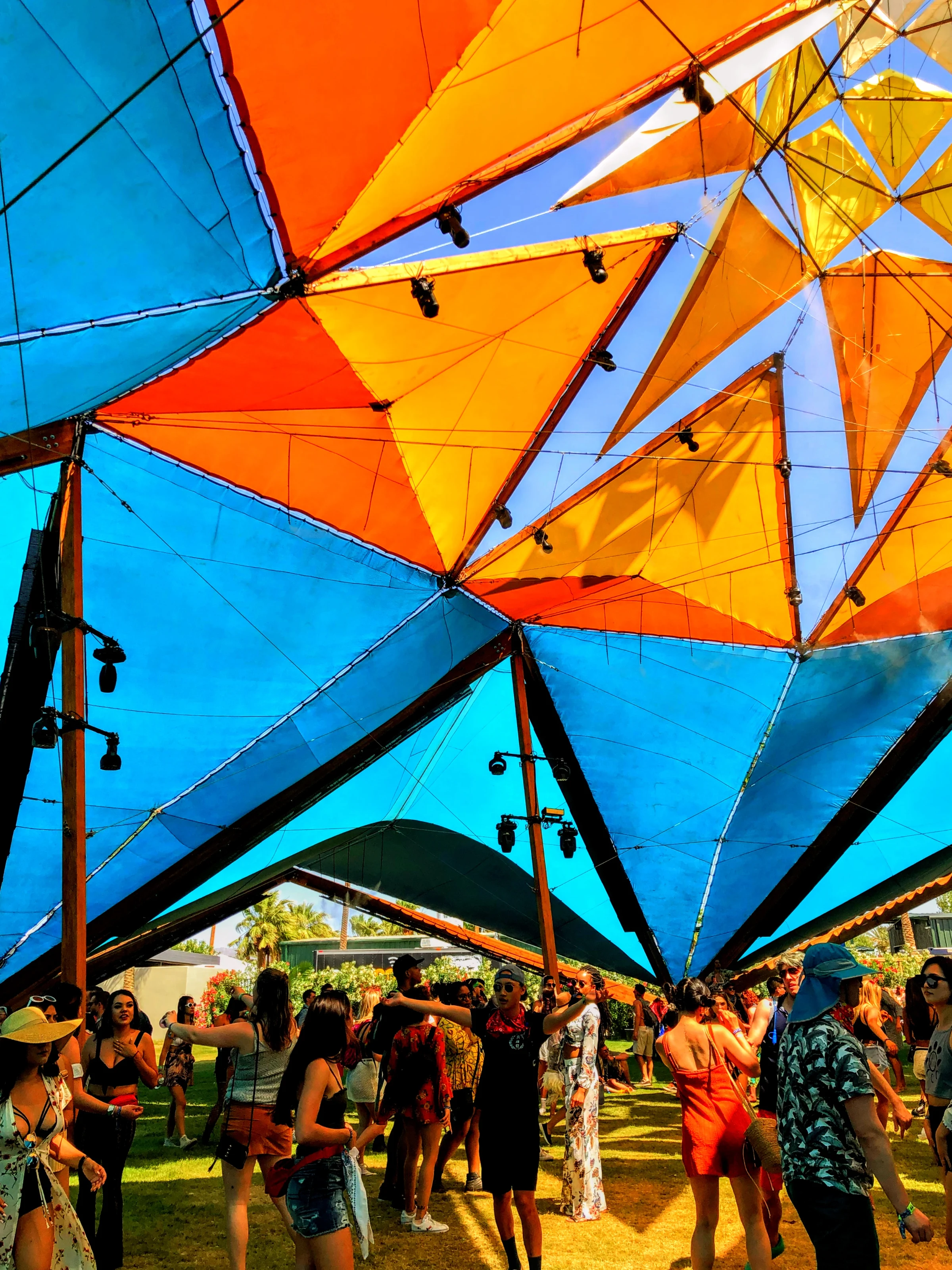 A crowd gathers on a grassy lawn beneath a geometric canopy constructed from vibrant blue, orange, and yellow triangular shade sails.