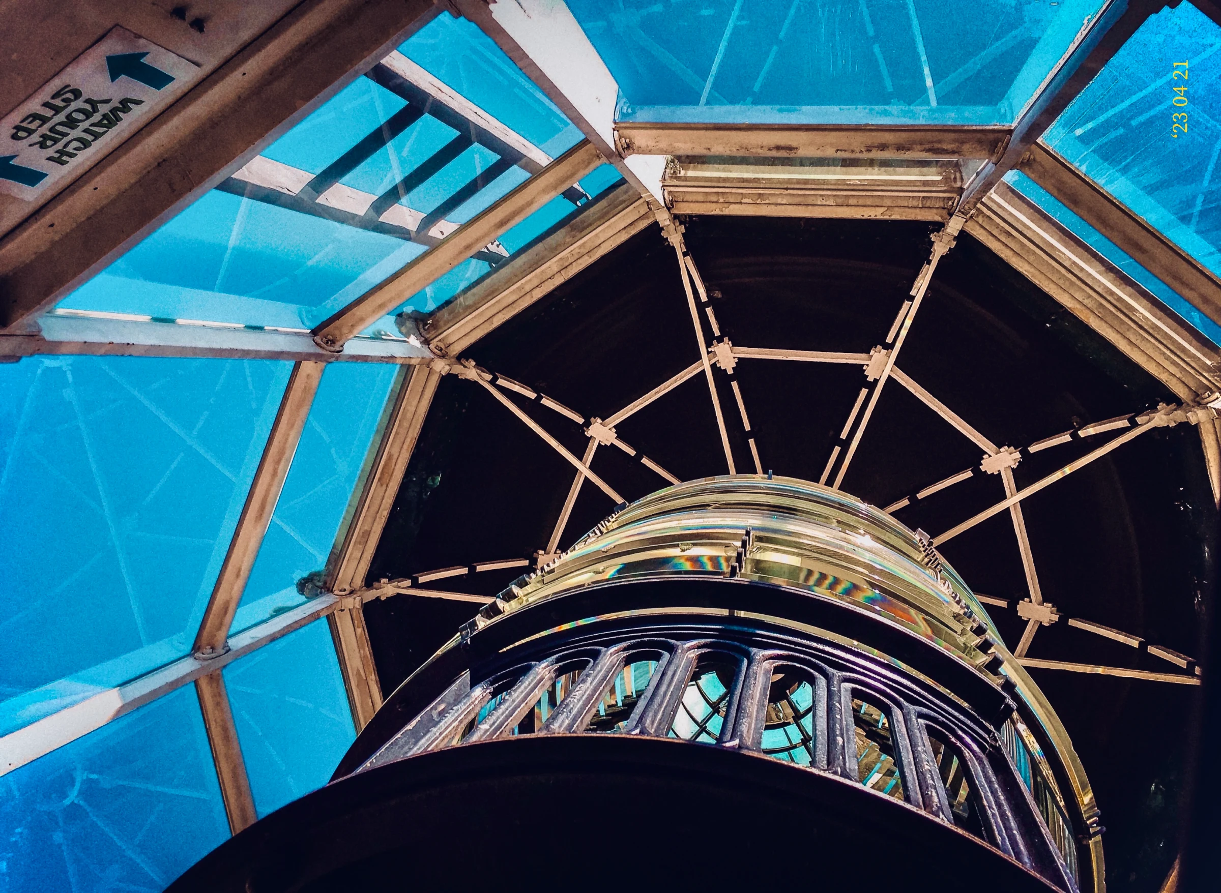 Low-angle view looking up inside a lighthouse lantern room, showcasing the glass Fresnel lens, dark iron framework, and blue glass panes.