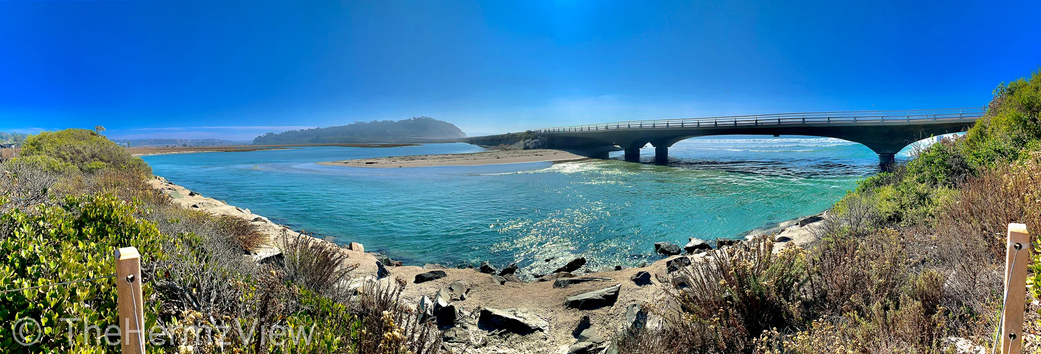 Panoramic view of a concrete bridge spanning a turquoise coastal inlet under a clear blue sky, framed by foreground scrub and wooden fence posts.
