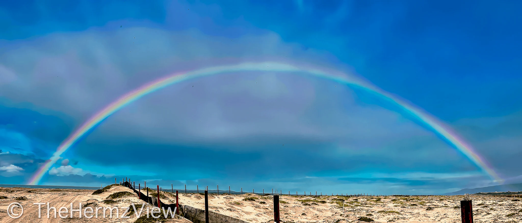 A full vibrant rainbow arcs across a bright blue sky over sandy dunes bordered by wooden fence posts.
