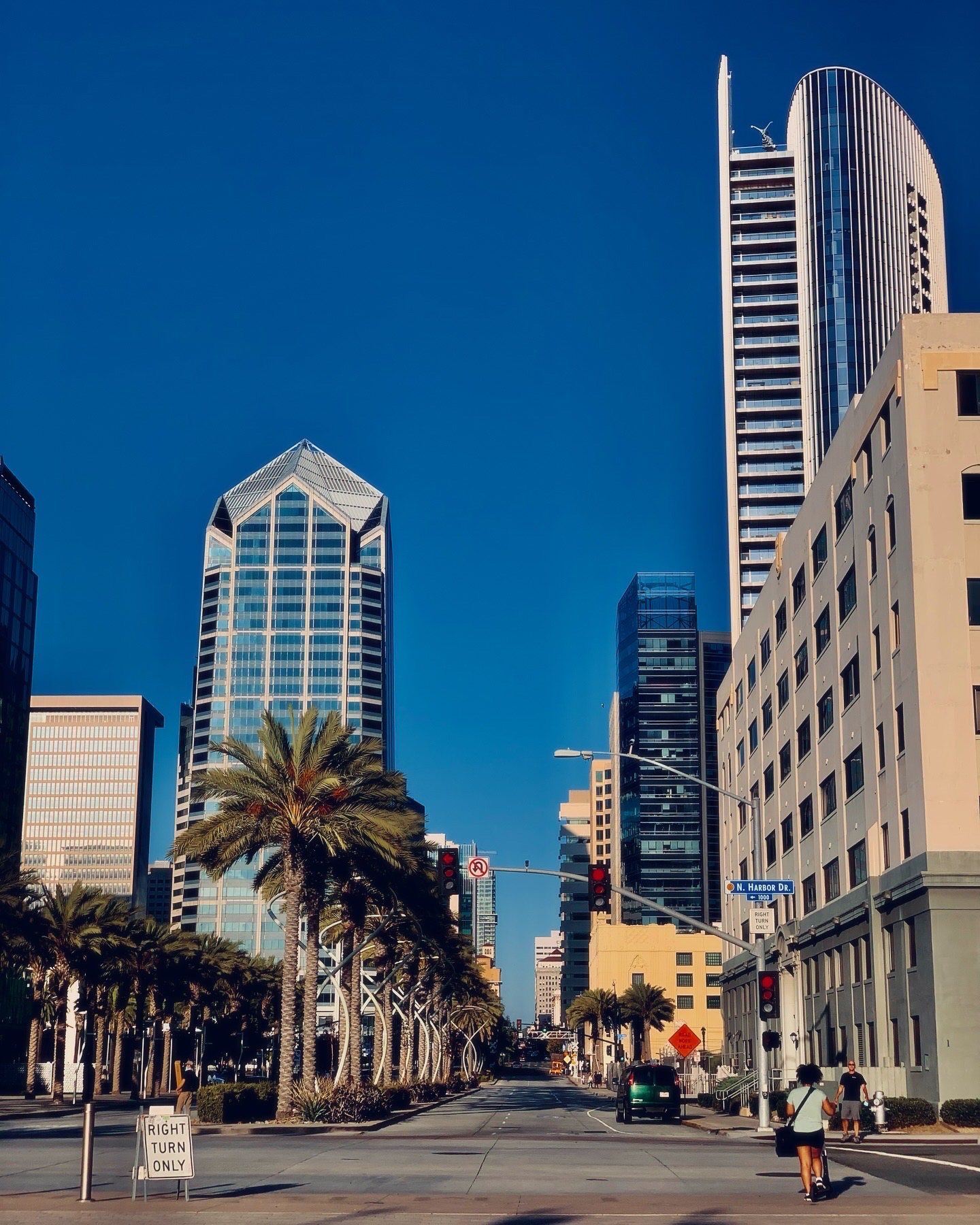 A wide urban avenue flanked by tall glass skyscrapers and palm trees under a clear blue sky.