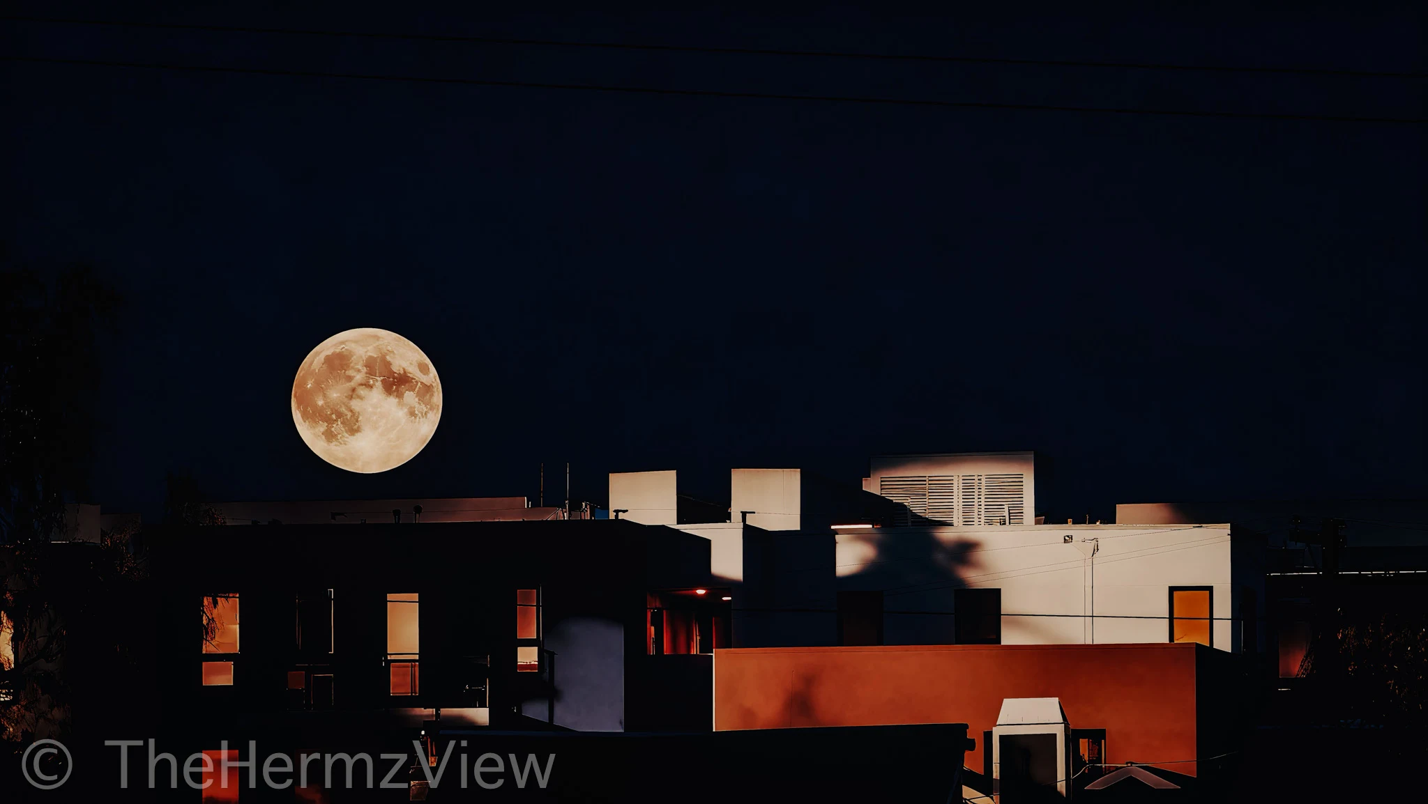 A detailed full moon low in a clear dark sky above modern building rooftops with illuminated windows.